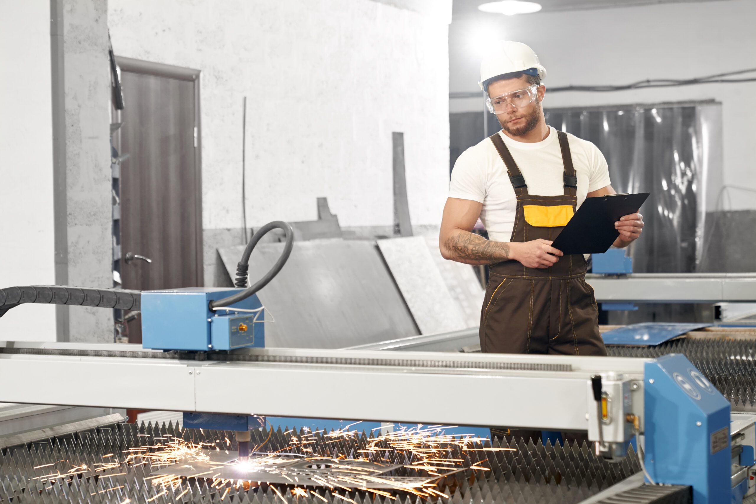 View from side of serious man in protective glasses and helmet keeping folder and writing down data while controlling process of devices and equipment. Concept of plasma cutting and metalwork.